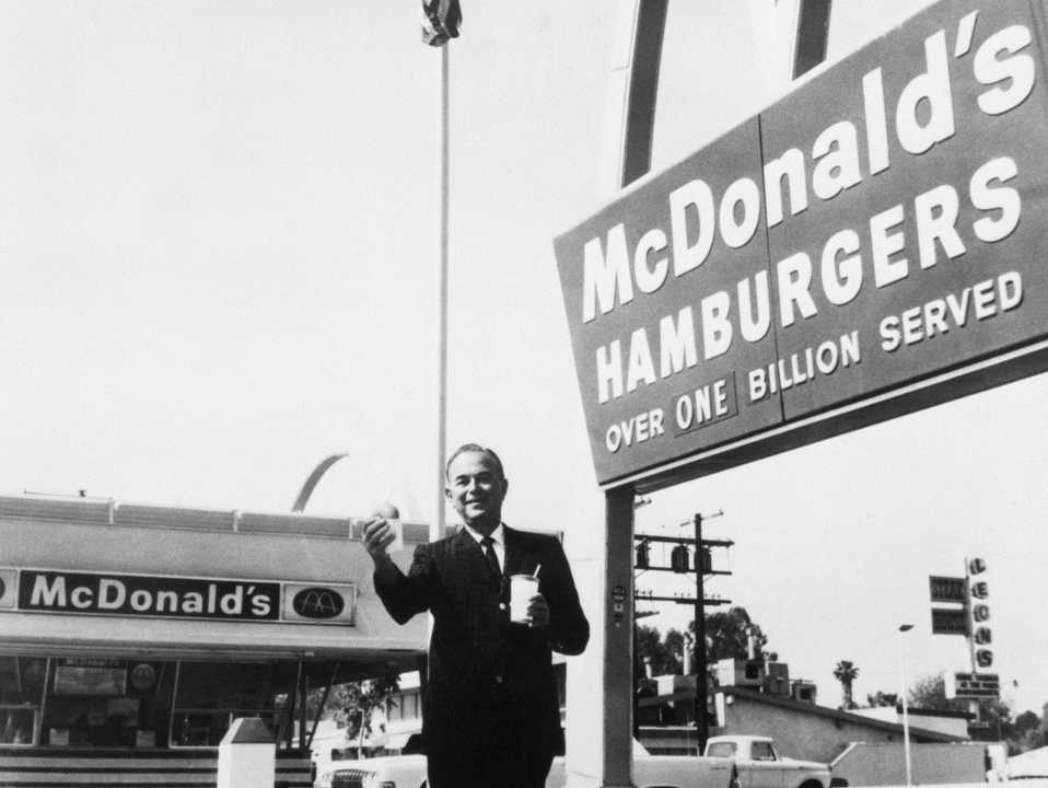 Founder and chairman of McDonald's Corporation, Ray Kroc, stands outside one of his franchises, holding a hamburger and a drink.