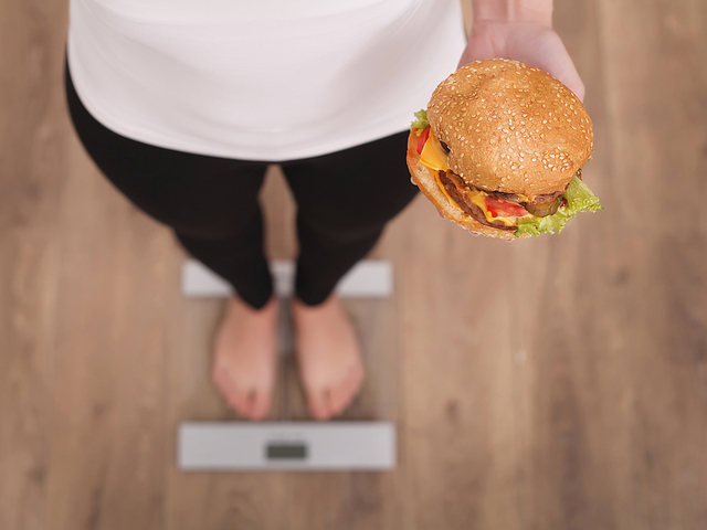 ハンバーガーを持ちながら体重計に乗る女性
